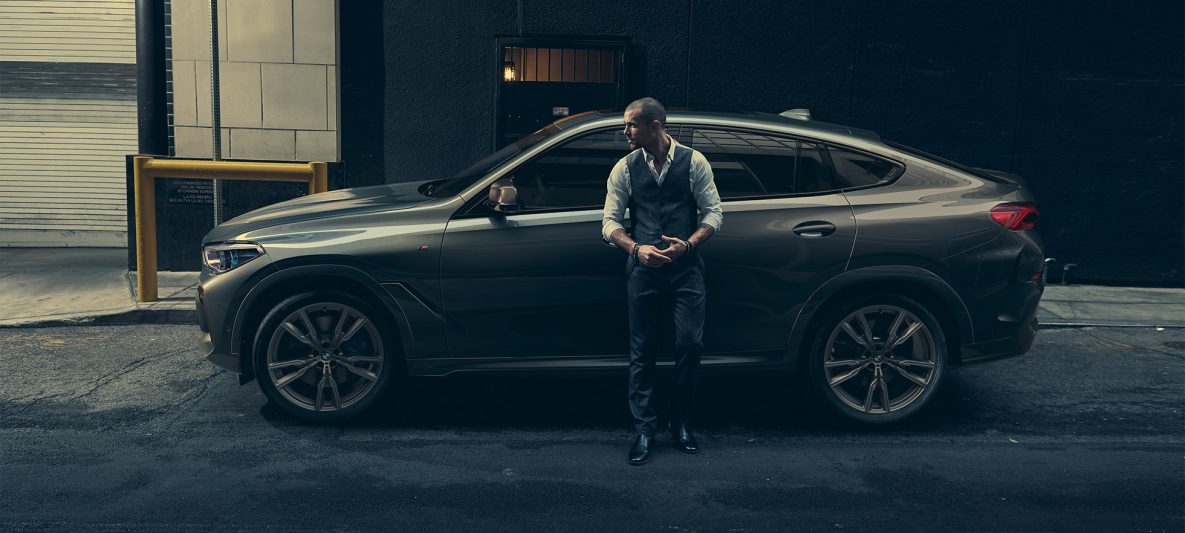 Mann lehnt an BMW X6 M50i in seitlicher Perspektive vor urbaner Kulisse